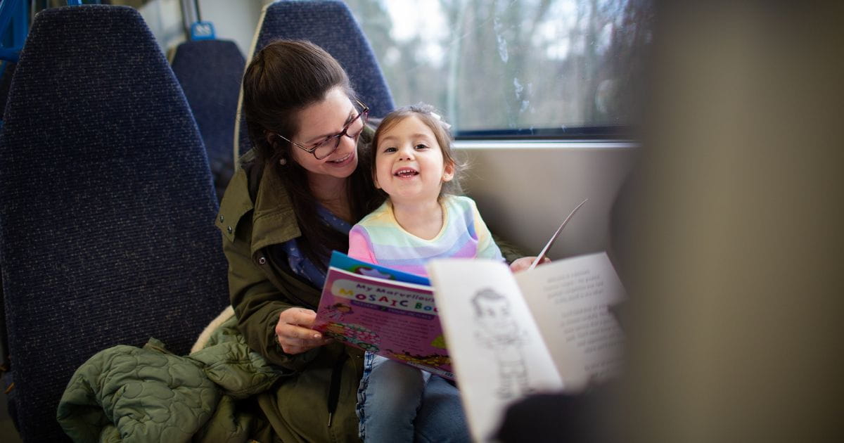 A woman with a child on her lap smiling and reading a book on the train